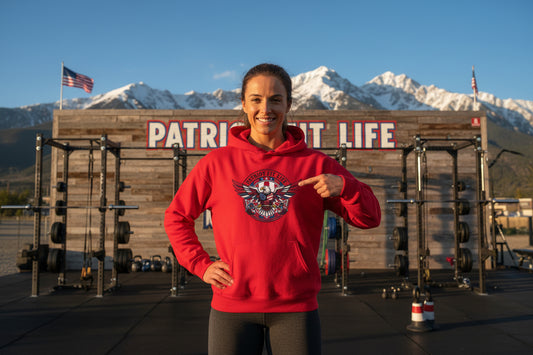 A woman wearing a Patriot Fit Life red hoodie with a logo, pointing to it, in front of a wooden building with a Patriot Fit Life sign and mountains.